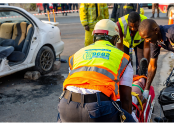 Emergency responders in high-visibility vests assist a person on a stretcher at the scene of a car crash nearby another damaged white car and debris on the road.
