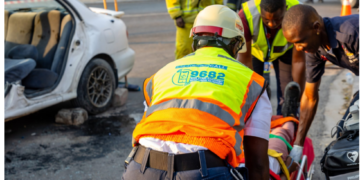 Emergency responders in high-visibility vests assist a person on a stretcher at the scene of a car crash nearby another damaged white car and debris on the road.