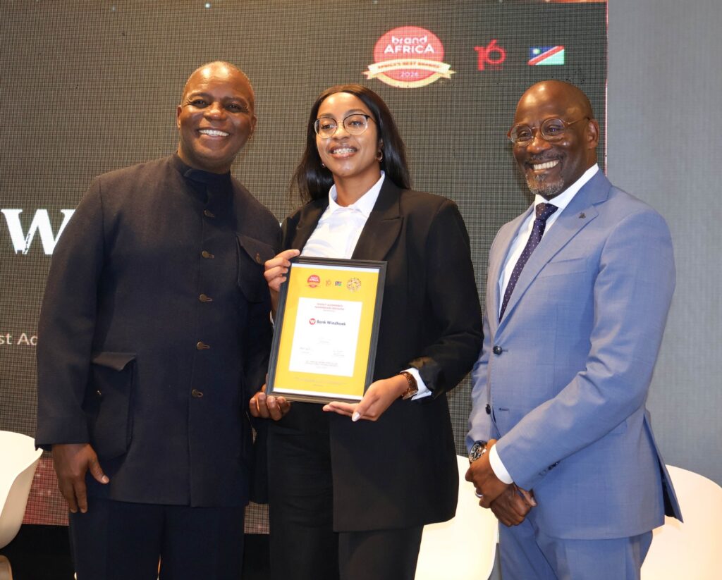 bwindhoek - The Brief | Namibia's Leading Business & Financial News Three people posing on stage; a woman in a black suit holds a framed award while flanked by two men in dark and light suits, all smiling.