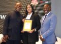bwindhoek - The Brief | Namibia's Leading Business & Financial News Three people posing on stage; a woman in a black suit holds a framed award while flanked by two men in dark and light suits, all smiling.