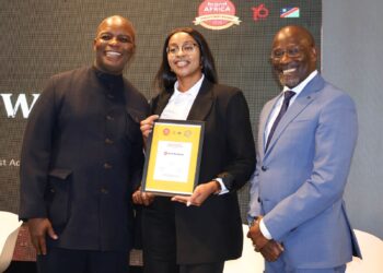 Three people posing on stage; a woman in a black suit holds a framed award while flanked by two men in dark and light suits, all smiling.