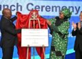 Two officials unveil a plaque on stage at a launch ceremony, with a woman in a green patterned outfit helping to remove the cloth.