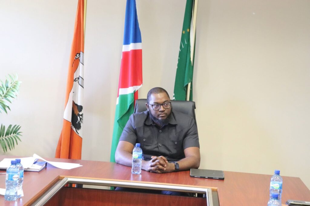 SAM NUJOMA - The Brief | Namibia's Leading Business & Financial News Man with glasses and beard seated at a conference table, with flags behind him in a meeting room.