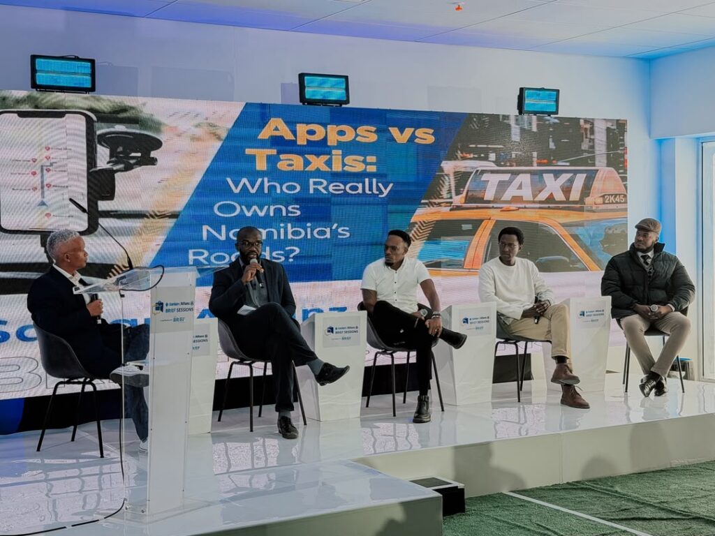 Panel discussion on a stage with five men seated; large screen behind reads 'Apps vs Taxis: Who Really Owns Namibia's Roads?'