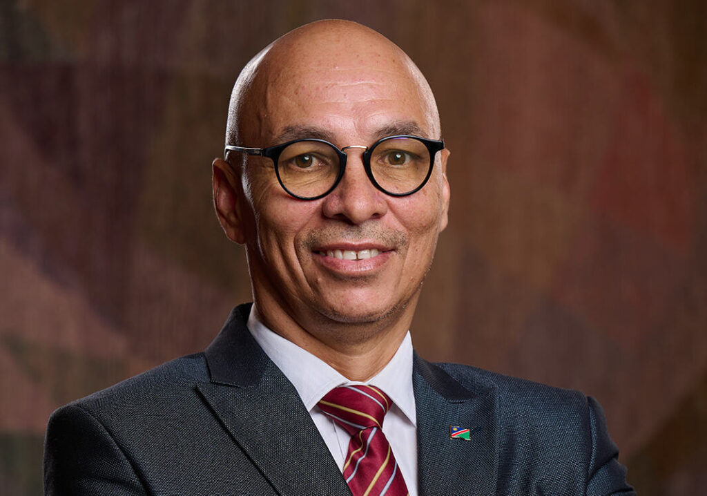 Head-and-shoulders portrait of a smiling man in a dark suit, white shirt, red striped tie, wearing round glasses and a flag lapel pin.