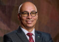 Head-and-shoulders portrait of a smiling man in a dark suit, white shirt, red striped tie, wearing round glasses and a flag lapel pin.