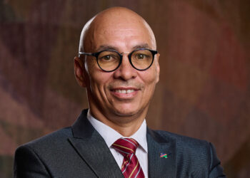 Head-and-shoulders portrait of a smiling man in a dark suit, white shirt, red striped tie, wearing round glasses and a flag lapel pin.