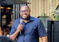 Khomas Gavoner - The Brief | Namibia's Leading Business & Financial News Smiling man in a navy short-sleeve shirt holding a microphone and speaking at an outdoor event.