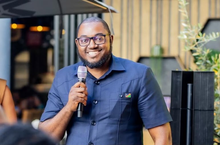 Khomas Gavoner - The Brief | Namibia's Leading Business & Financial News Smiling man in a navy short-sleeve shirt holding a microphone and speaking at an outdoor event.