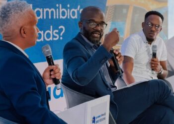 Panel of three men in suits on stage, each holding a microphone; blue banner behind reads 'Who Really Owns ...' with taxi imagery in the background.