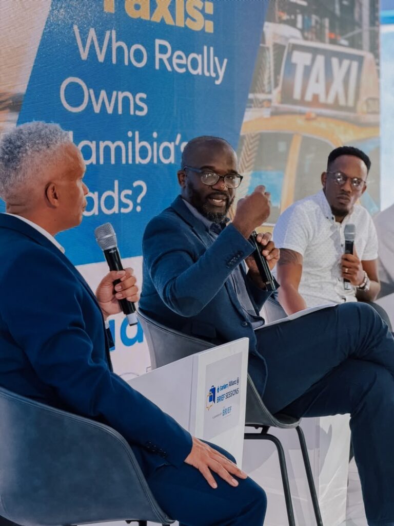 gov public transport - The Brief | Namibia's Leading Business & Financial News Panel of three men in suits on stage, each holding a microphone; blue banner behind reads 'Who Really Owns ...' with taxi imagery in the background.