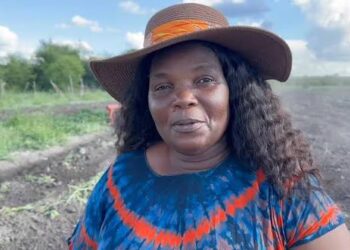 Portrait of a woman wearing a wide straw hat and a blue-red tie-dye shirt, standing in a dirt field with greenery in the background.