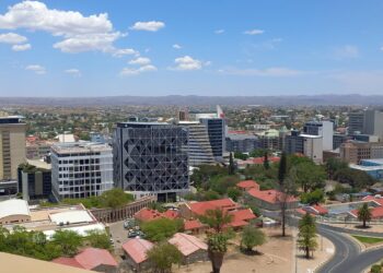 City skyline with modern glass buildings, red-roofed houses in foreground, and distant hills under a clear blue sky.