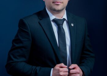 Professional head-and-shoulders portrait of a man in a dark suit and tie against a blue gradient background.