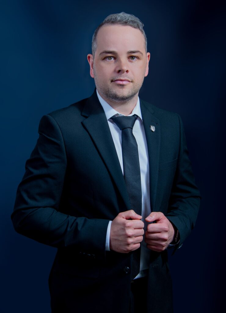 Professional head-and-shoulders portrait of a man in a dark suit and tie against a blue gradient background.