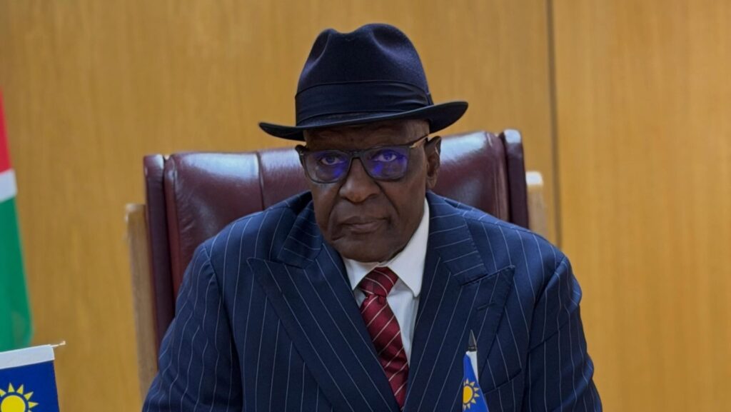 KAIRE MBUENDE5 - The Brief | Namibia's Leading Business & Financial News Older man in a navy pinstripe suit with a red tie, black hat and glasses, seated at a desk in a formal setting.