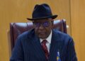 KAIRE MBUENDE5 - The Brief | Namibia's Leading Business & Financial News Older man in a navy pinstripe suit with a red tie, black hat and glasses, seated at a desk in a formal setting.