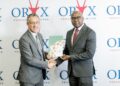 Two businessmen in gray suits shake hands as one passes a green folder at a corporate event with ORX logos behind them behind the backdrop.