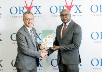 Two businessmen in gray suits shake hands as one passes a green folder at a corporate event with ORX logos behind them behind the backdrop.