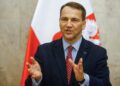 Radosław Sikorski - The Brief | Namibia's Leading Business & Financial News Man in a dark suit and striped shirt speaking in front of red-and-white flags, using hand gestures for emphasis.
