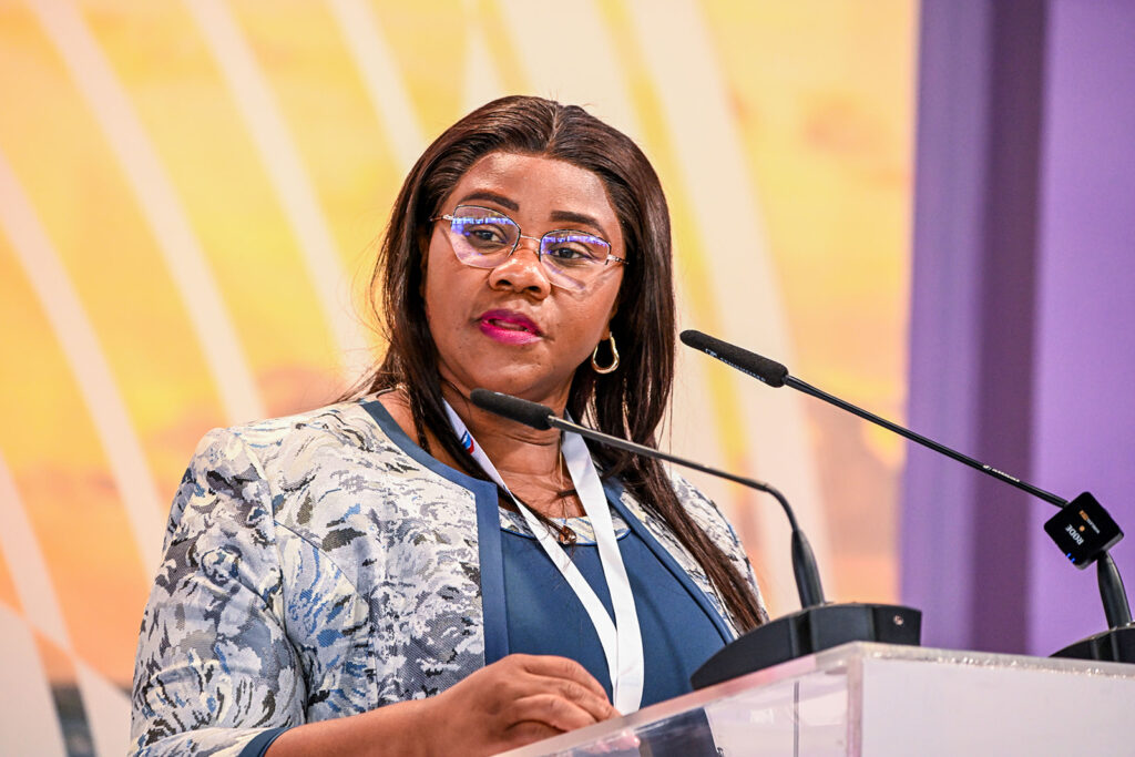 Woman in a blue blouse and patterned jacket speaks at a podium with two microphones at a conference event.
