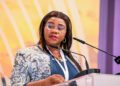 VICTORIA SIBEYA 2 - The Brief | Namibia's Leading Business & Financial News Woman in a blue blouse and patterned jacket speaks at a podium with two microphones at a conference event.