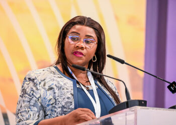 Woman in a blue blouse and patterned jacket speaks at a podium with two microphones at a conference event.