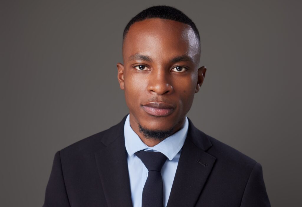 Portrait of a young man in a dark suit and blue tie against a gray background.