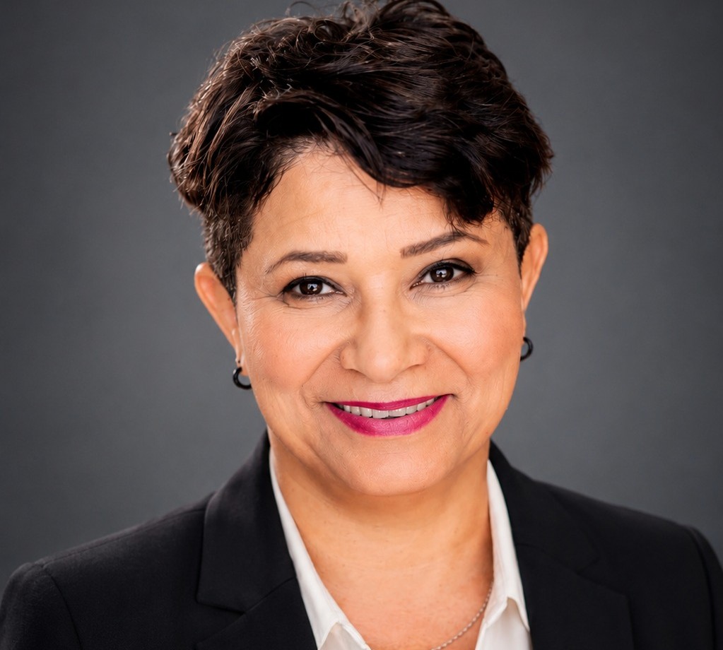 Professional headshot of a smiling woman in a black blazer against a gray backdrop.