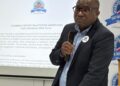 Fluksman Samuehl2 - The Brief | Namibia's Leading Business & Financial News Male presenter in a dark suit speaks into a handheld microphone in front of a slide about the Public Enterprises CEOs Forum, with logos on the screen and banner nearby.