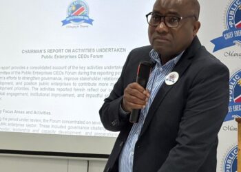 Male presenter in a dark suit speaks into a handheld microphone in front of a slide about the Public Enterprises CEOs Forum, with logos on the screen and banner nearby.