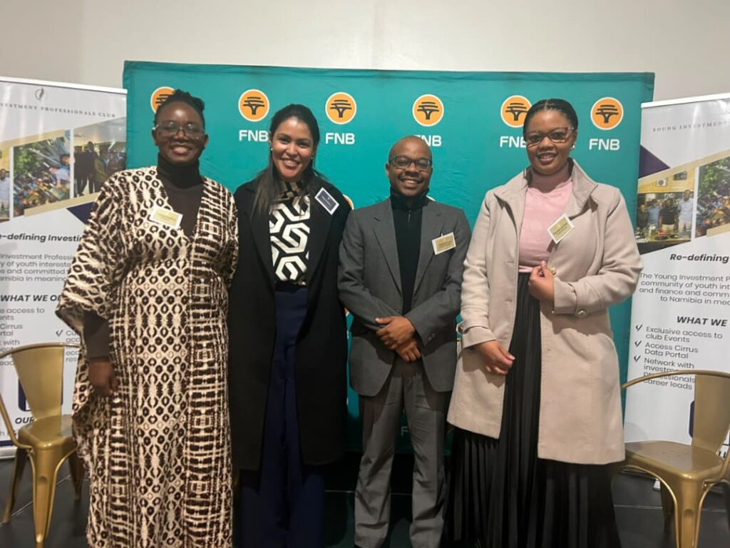 Four professionals posing and smiling at a corporate event with FNB banners in turquoise behind them, name tags visible.