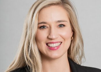 Smiling woman with long blonde hair wearing a black blazer against a gray background (professional headshot).