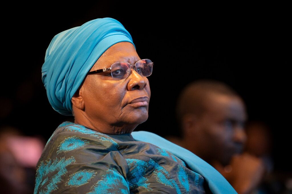 NETUMBO 8 - The Brief | Namibia's Leading Business & Financial News Older woman wearing a turquoise headwrap and glasses, seated in a dimly lit audience.