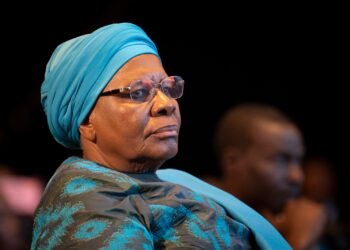 NETUMBO 8 - The Brief | Namibia's Leading Business & Financial News Older woman wearing a turquoise headwrap and glasses, seated in a dimly lit audience.