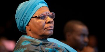 Older woman wearing a turquoise headwrap and glasses, seated in a dimly lit audience.