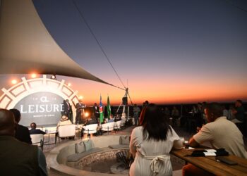 Rooftop evening event with a stage, large circular ‘Leisure’ backdrop, and audience seated around tables at sunset.