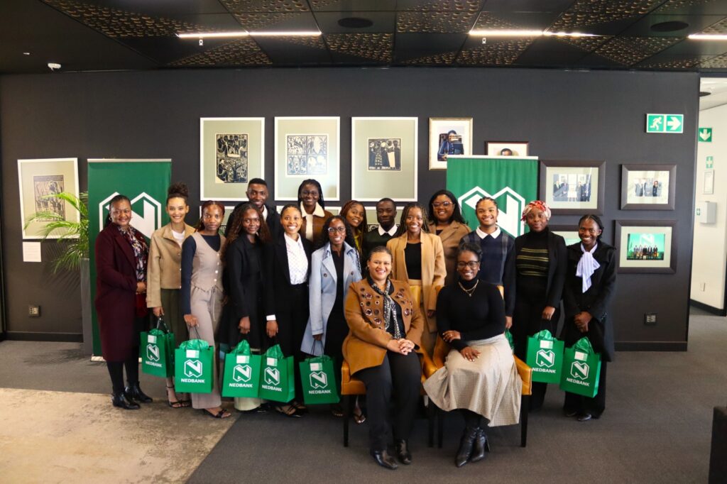Group of people posing for a photo at an event, holding green Nedbank gift bags in a gallery-like setting with framed art on dark walls.