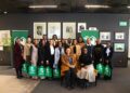 Group of people posing for a photo at an event, holding green Nedbank gift bags in a gallery-like setting with framed art on dark walls.
