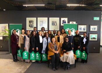 Group of people posing for a photo at an event, holding green Nedbank gift bags in a gallery-like setting with framed art on dark walls.