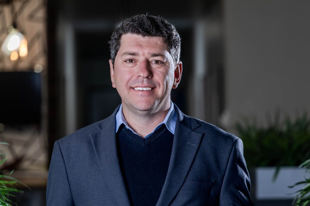 Smiling man in a navy blazer and light blue shirt standing in a modern indoor setting