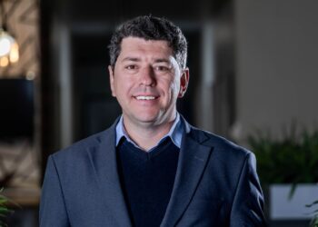 Smiling man in a navy blazer and light blue shirt standing in a modern indoor setting