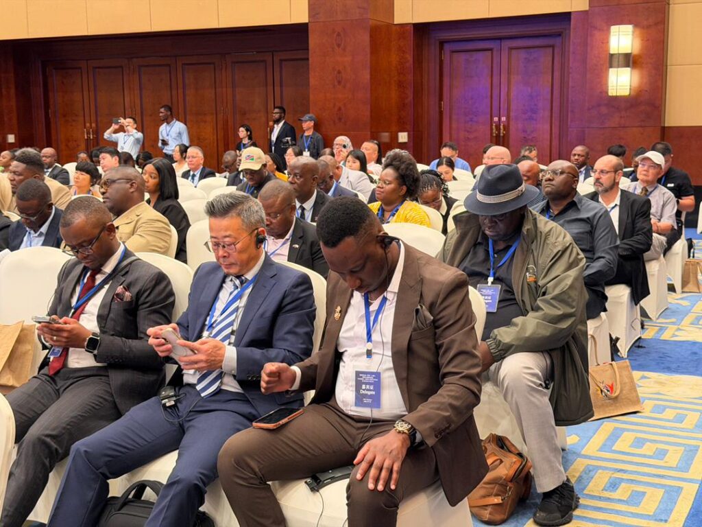 Audience of diverse professionals seated in a conference hall, some looking at phones, wearing lanyards and badges at a formal event.