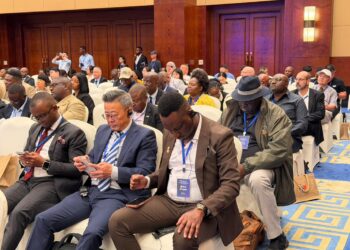 Audience of diverse professionals seated in a conference hall, some looking at phones, wearing lanyards and badges at a formal event.