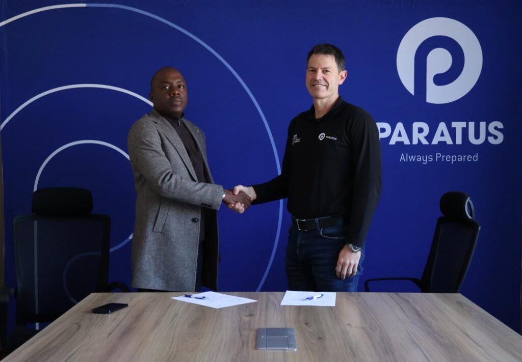 Two men shake hands across a conference table in front of a blue wall with the Paratus logo, signifying a business agreement.
