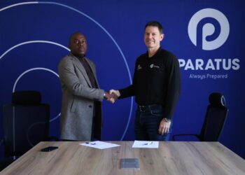 Two men shake hands across a conference table in front of a blue wall with the Paratus logo, signifying a business agreement.