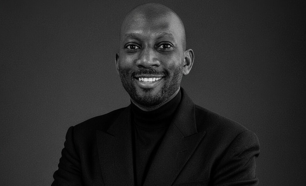 Smiling man in a dark blazer and black turtleneck posing for a professional portrait against a dark gray background.