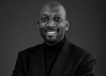 Smiling man in a dark blazer and black turtleneck posing for a professional portrait against a dark gray background.