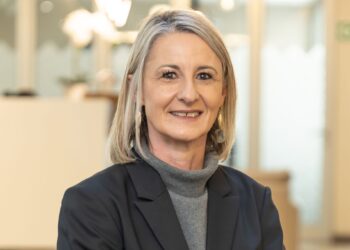 Portrait of a smiling blonde woman in a gray turtleneck and dark blazer, in an office setting behind her.