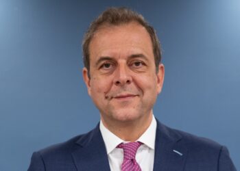 Professional headshot of a smiling man in a navy suit and pink tie against a blue background.